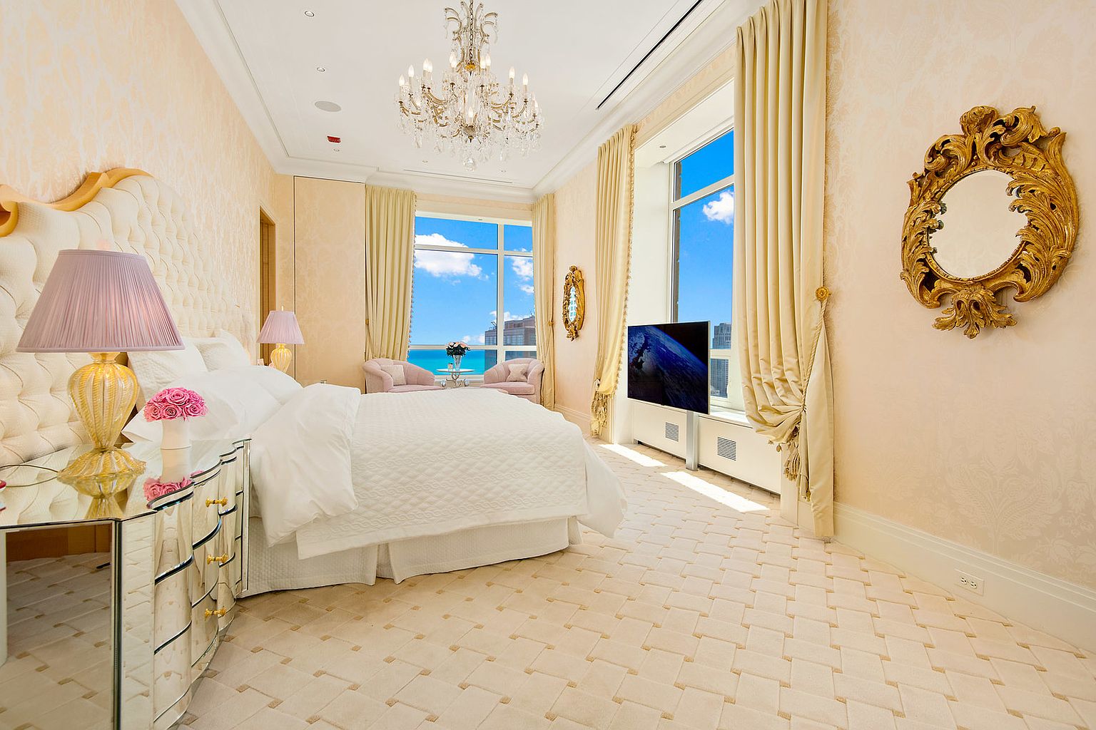 This is a luxurious primary bedroom featuring a large bed with a tufted headboard and white bedding. The room is decorated in a soft, elegant style with patterned wallpaper, a crystal chandelier, and floor-to-ceiling windows offering a view of the city and ocean. A gilded mirror and mirrored nightstand add to the room's opulence.