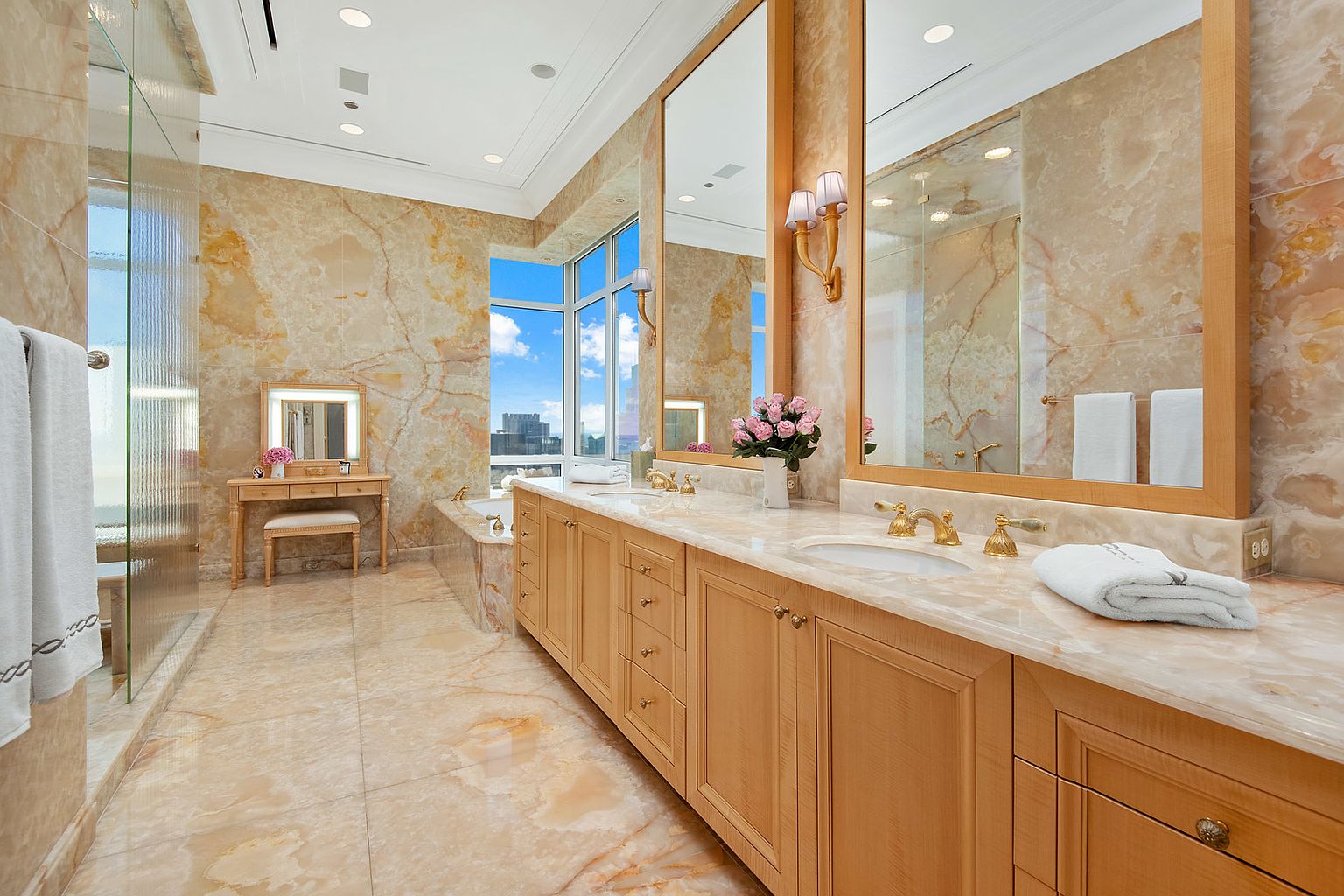 This is a luxurious primary bathroom featuring marble walls and flooring, a long vanity with dual sinks, and large mirrors. The bathroom also includes a glass-enclosed shower and a separate vanity area with a stool. The overall impression is elegant and upscale, with a focus on high-end finishes and ample natural light from the large window.