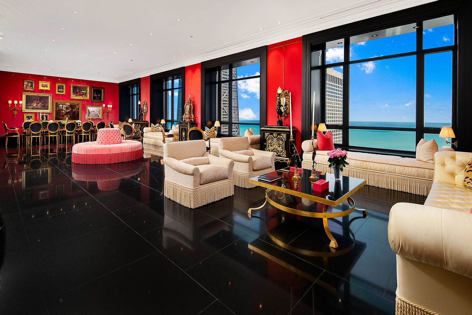 This is a luxurious living room featuring a glossy black tile floor, red accent walls, and large windows offering a stunning view of the city and lake. The room is furnished with elegant sofas, armchairs with fringe, and a glass-topped coffee table with gold accents. A formal dining area is visible in the background, creating a sense of grandeur and sophistication.