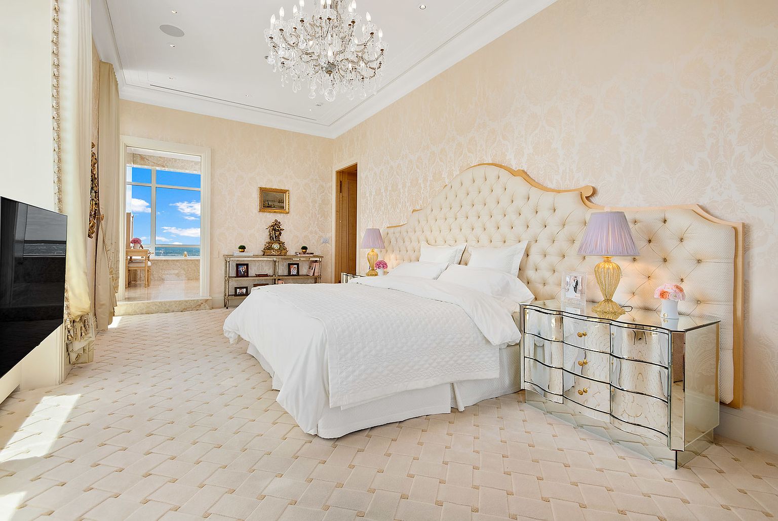 This is a luxurious primary bedroom featuring a large, tufted headboard and crisp white bedding, creating a serene and inviting atmosphere. Mirrored nightstands flank the bed, adding a touch of glamour, while a crystal chandelier illuminates the room. The room opens to a balcony with ocean views, enhancing the sense of opulence and relaxation.