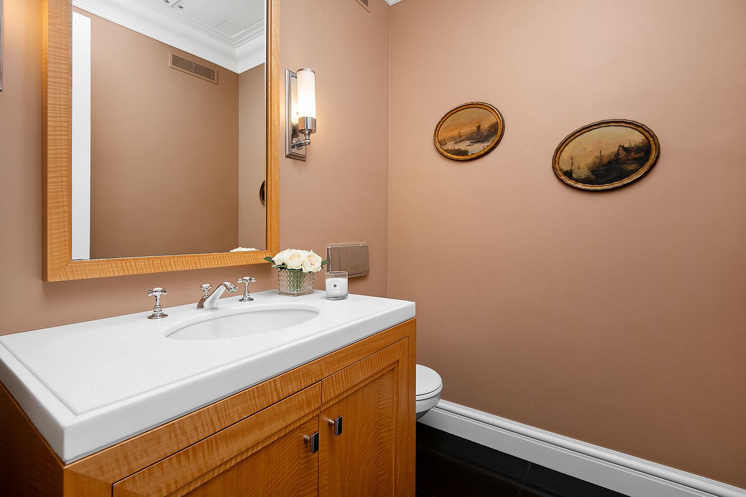 This is a well-appointed guest bathroom featuring a wooden vanity with a white countertop and an oval sink. A framed mirror hangs above the sink, complemented by sconce lighting. Two framed paintings adorn the wall, adding a touch of elegance to the space.