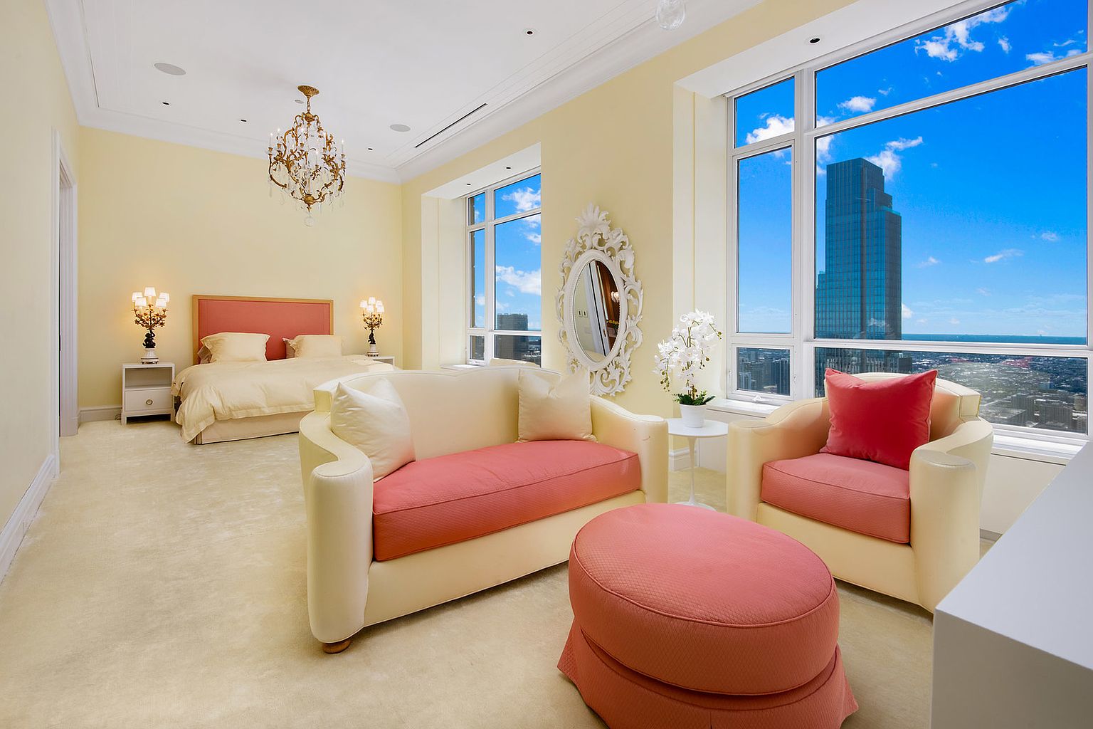 This primary bedroom features a luxurious and spacious layout with a seating area. The room is decorated in soft, warm tones, with a large bed, a stylish sofa, an armchair, and an ottoman. Large windows offer stunning city views, enhancing the room's appeal.