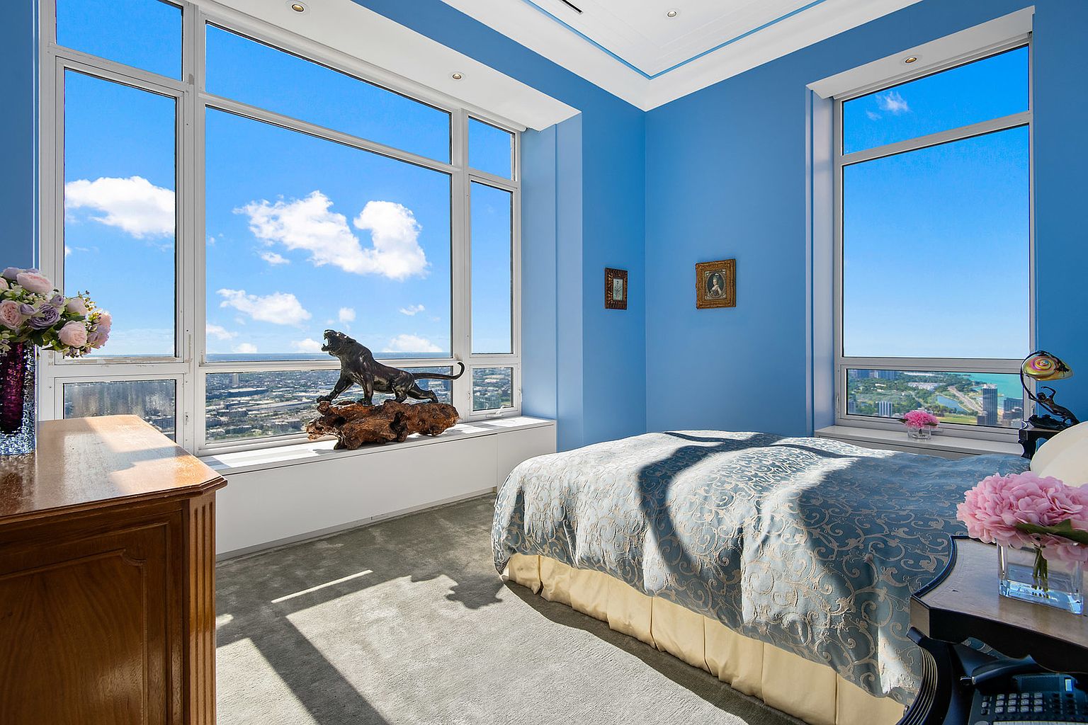 This is a primary bedroom with blue walls and large windows offering a city view. The room features a bed with a patterned blue and gold comforter, a wooden dresser, and decorative items like a lion sculpture on the windowsill and floral arrangements. The overall impression is luxurious and bright, emphasizing the stunning cityscape.