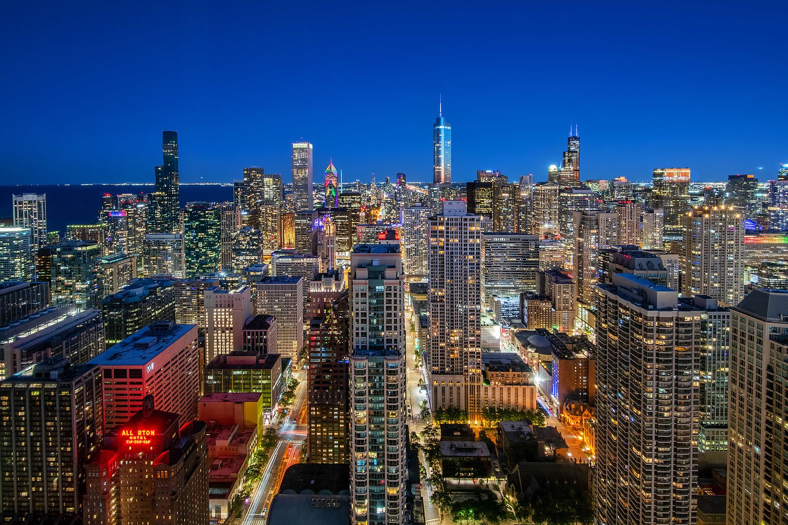 This aerial view showcases the stunning Chicago skyline at night, with numerous illuminated skyscrapers creating a vibrant urban landscape. The image captures the density and grandeur of the city, highlighting its architectural diversity and the bustling atmosphere. The deep blue sky provides a dramatic backdrop, emphasizing the city's impressive scale and beauty.