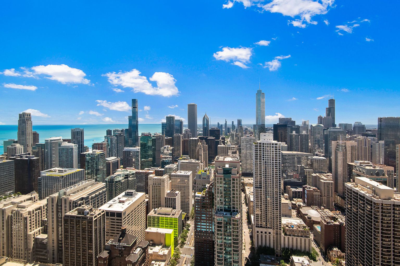 This aerial view showcases a dense urban cityscape under a bright blue sky dotted with fluffy white clouds. Numerous skyscrapers of varying heights and architectural styles dominate the scene, creating a dynamic skyline. The image captures the scale and vibrancy of the city, emphasizing its modern architecture and bustling atmosphere.