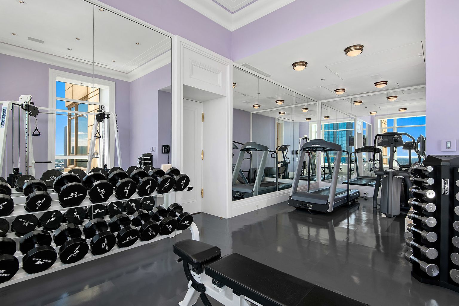 This is an interior shot of a home gym featuring a variety of exercise equipment, including dumbbells, a weight machine, and a treadmill. The room has a mirrored wall that enhances the sense of space and reflects the equipment, while the gray flooring and light purple walls create a modern and clean aesthetic. The presence of natural light from the windows adds to the appeal of the workout space.