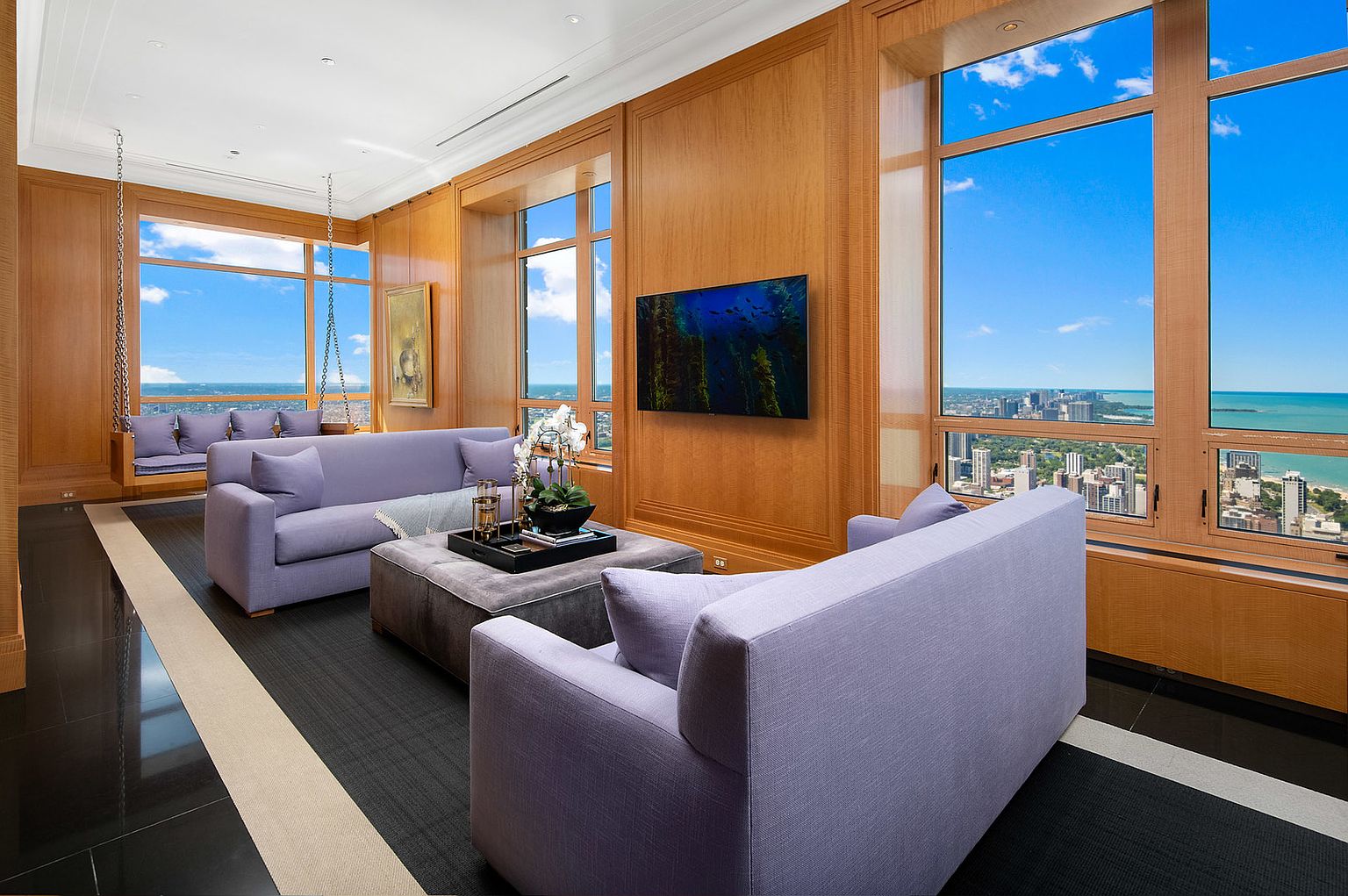 This is a luxurious living room featuring wood paneled walls, large windows with city and lake views, and stylish furniture. The room includes two lavender sofas, a gray ottoman, and a swing seat by the window. A flat-screen TV is mounted on the wall, and the flooring is a mix of black and gray with a light-colored stripe.