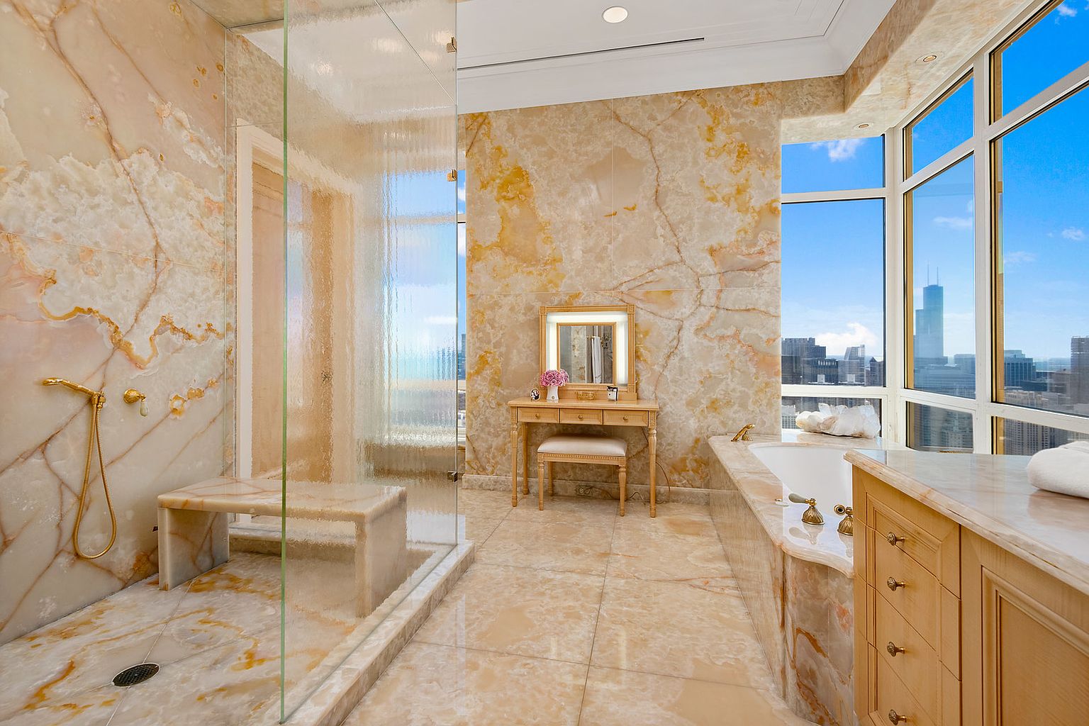 This is a luxurious primary bathroom featuring marble walls and flooring, a glass-enclosed shower with a built-in bench, and a vanity area with a lighted mirror. A soaking tub is positioned near large windows offering city views, enhancing the spa-like atmosphere. The space is well-lit and exudes elegance and sophistication.
