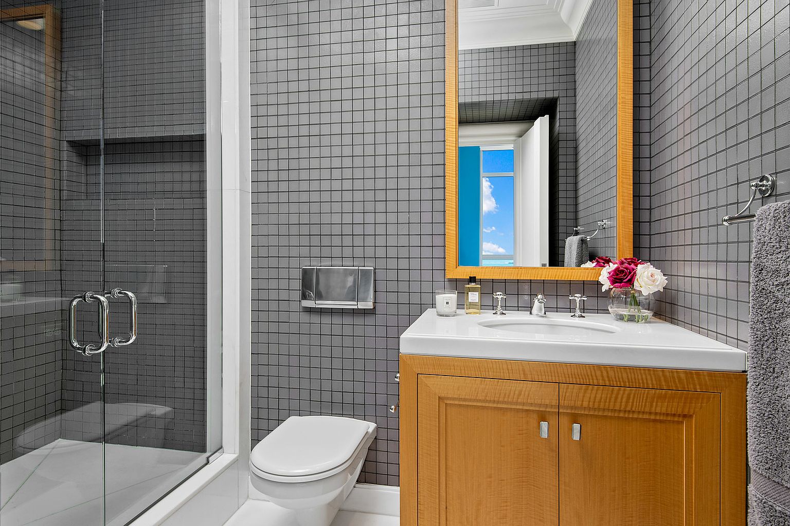 This is a well-appointed guest bathroom featuring gray tiled walls, a modern vanity with a white countertop, and a framed mirror. A glass-enclosed shower is visible to the left, and a toilet is positioned next to the vanity. The overall impression is clean, contemporary, and functional.