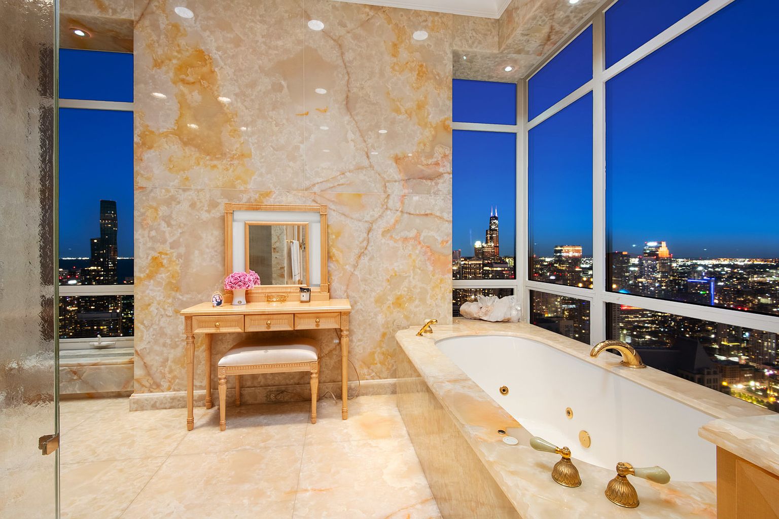 This is a luxurious primary bathroom featuring marble walls and flooring, a large soaking tub with gold fixtures, and a vanity with a mirror and stool. The bathroom has large windows offering a stunning city view at night. The perspective is from the doorway, showcasing the entire space.