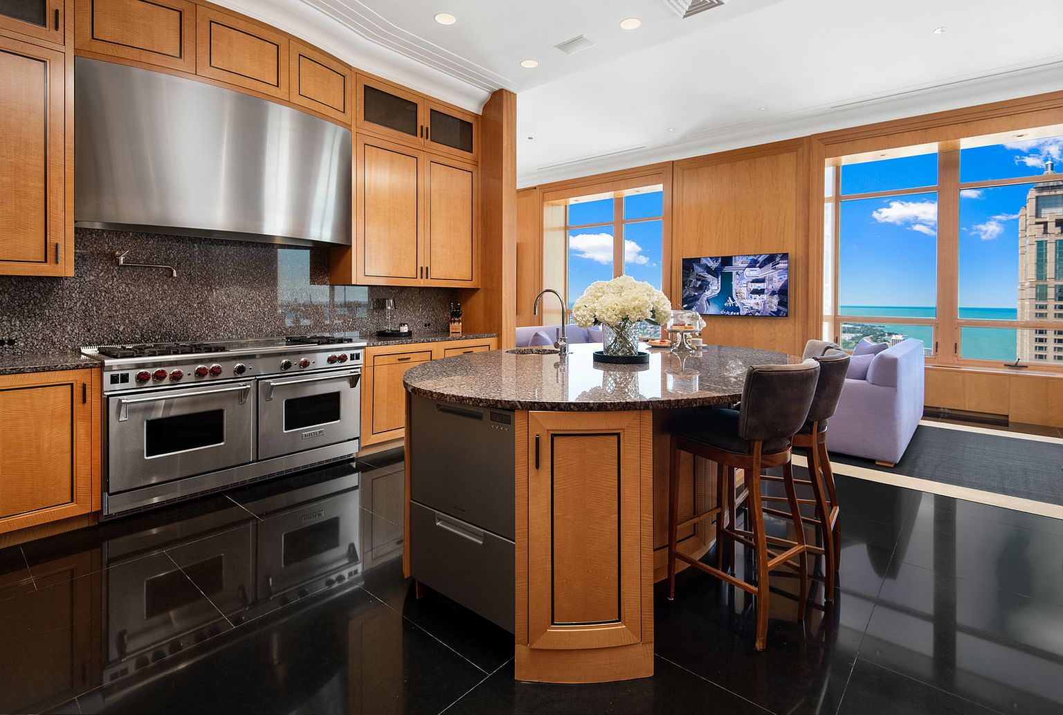 This is a luxurious kitchen featuring wood cabinetry, granite countertops, and stainless steel appliances, including a large Wolf range. A round island with bar seating anchors the space, while large windows offer stunning city and lake views. The black tile flooring adds a touch of elegance and reflects the natural light.