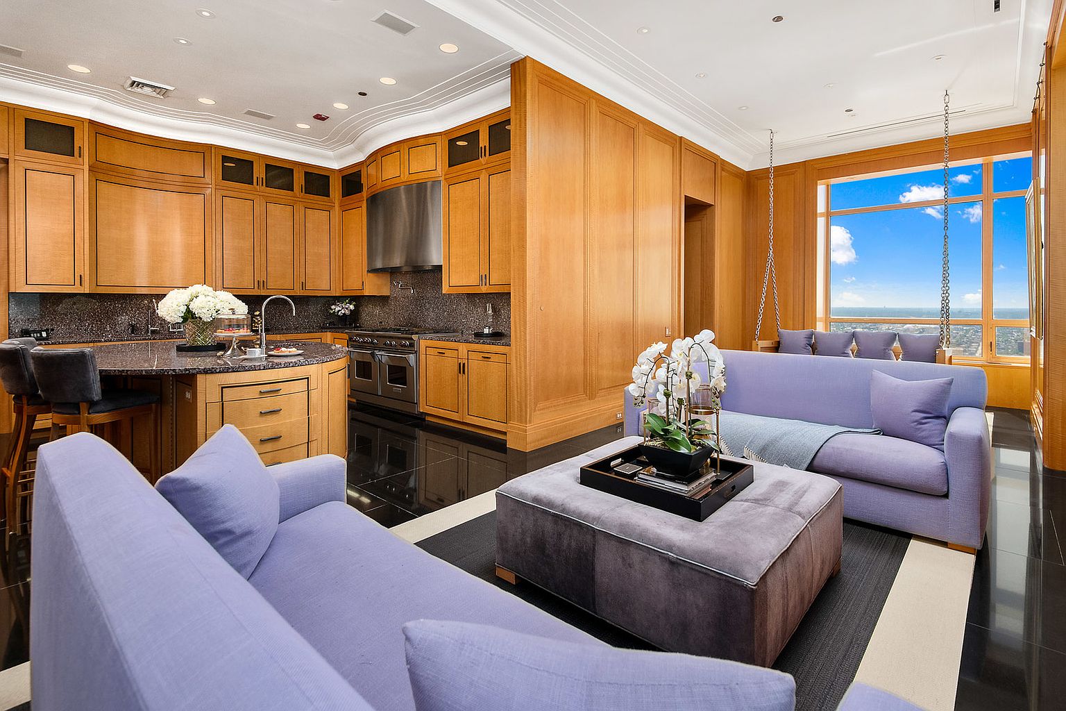 This interior shot showcases a luxurious living space that seamlessly integrates with a modern kitchen. The room features elegant wood paneling, a comfortable lavender sofa, and a unique swing chair by the window, offering stunning views. The black tile flooring adds a touch of sophistication, while the overall design exudes comfort and high-end living.