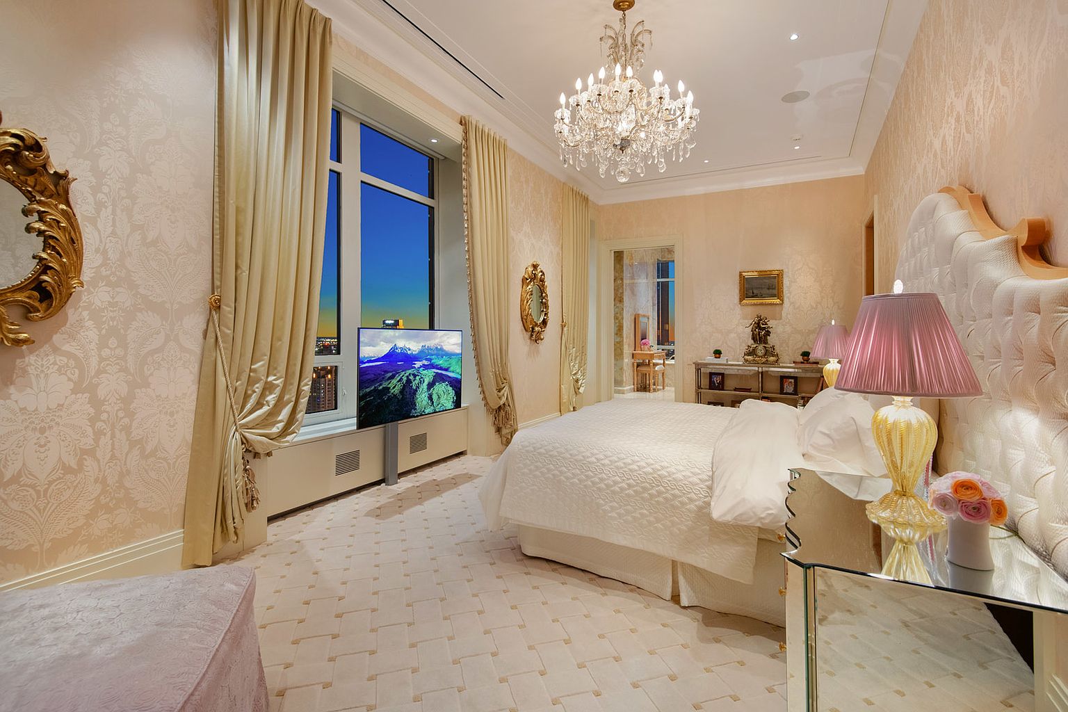 This is a luxurious primary bedroom featuring a large bed with a tufted headboard and white bedding. The room is decorated in a soft, elegant style with patterned wallpaper, a crystal chandelier, and floor-to-ceiling curtains. A large window offers a view of the city skyline, and a television is mounted in front of it.
