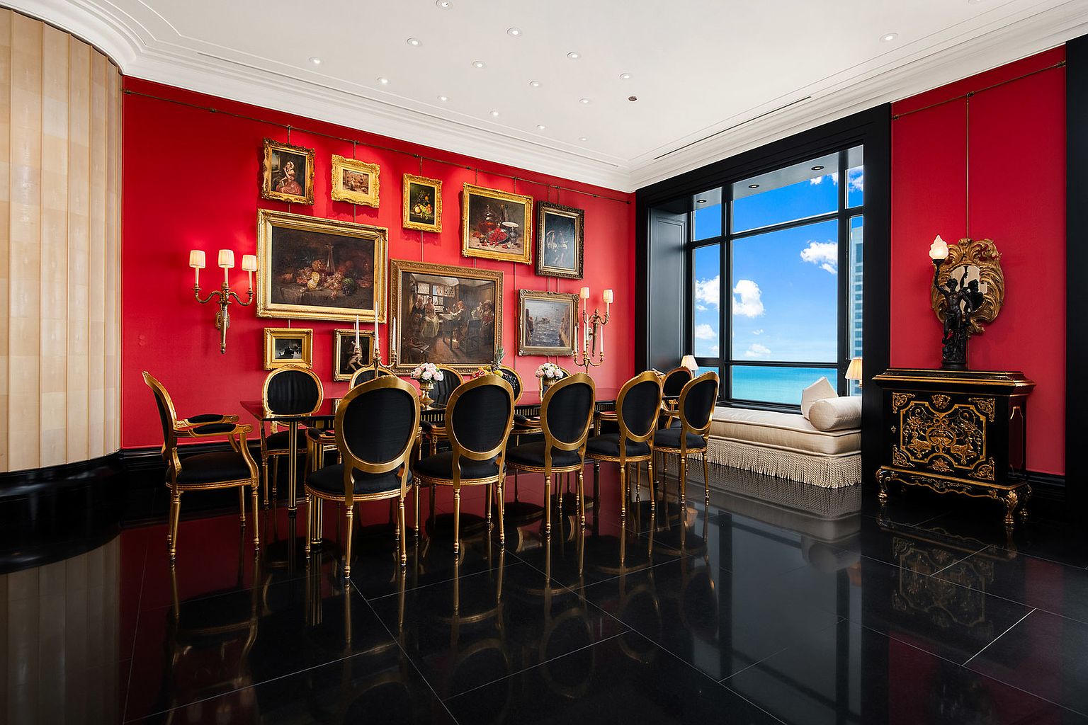 This is an opulent dining room featuring a red accent wall adorned with various framed artworks. A dark, glossy floor reflects the light from the recessed ceiling lights and the large window offering a view of the ocean. The dining set includes black chairs with gold frames, complementing the room's luxurious and classic style.