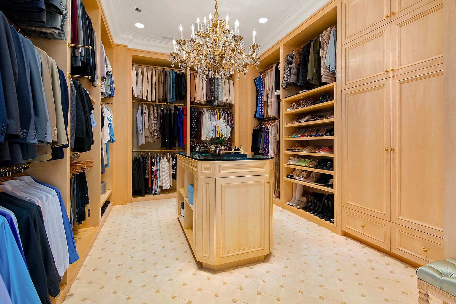 This is a luxurious walk-in closet featuring custom wood cabinetry, ample hanging space, and shelving for shoes and accessories. A central island with a dark countertop provides additional storage and display space. The room is illuminated by an ornate chandelier, creating an elegant and organized space.