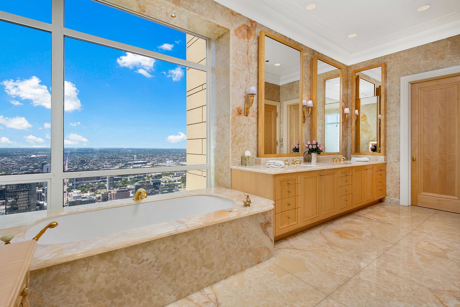 This is a luxurious primary bathroom featuring marble walls and flooring, a large soaking tub with gold fixtures positioned in front of a large window with a city view. The bathroom also includes a long vanity with three mirrors, sconces, and light wood cabinetry. The overall impression is one of opulence and relaxation.