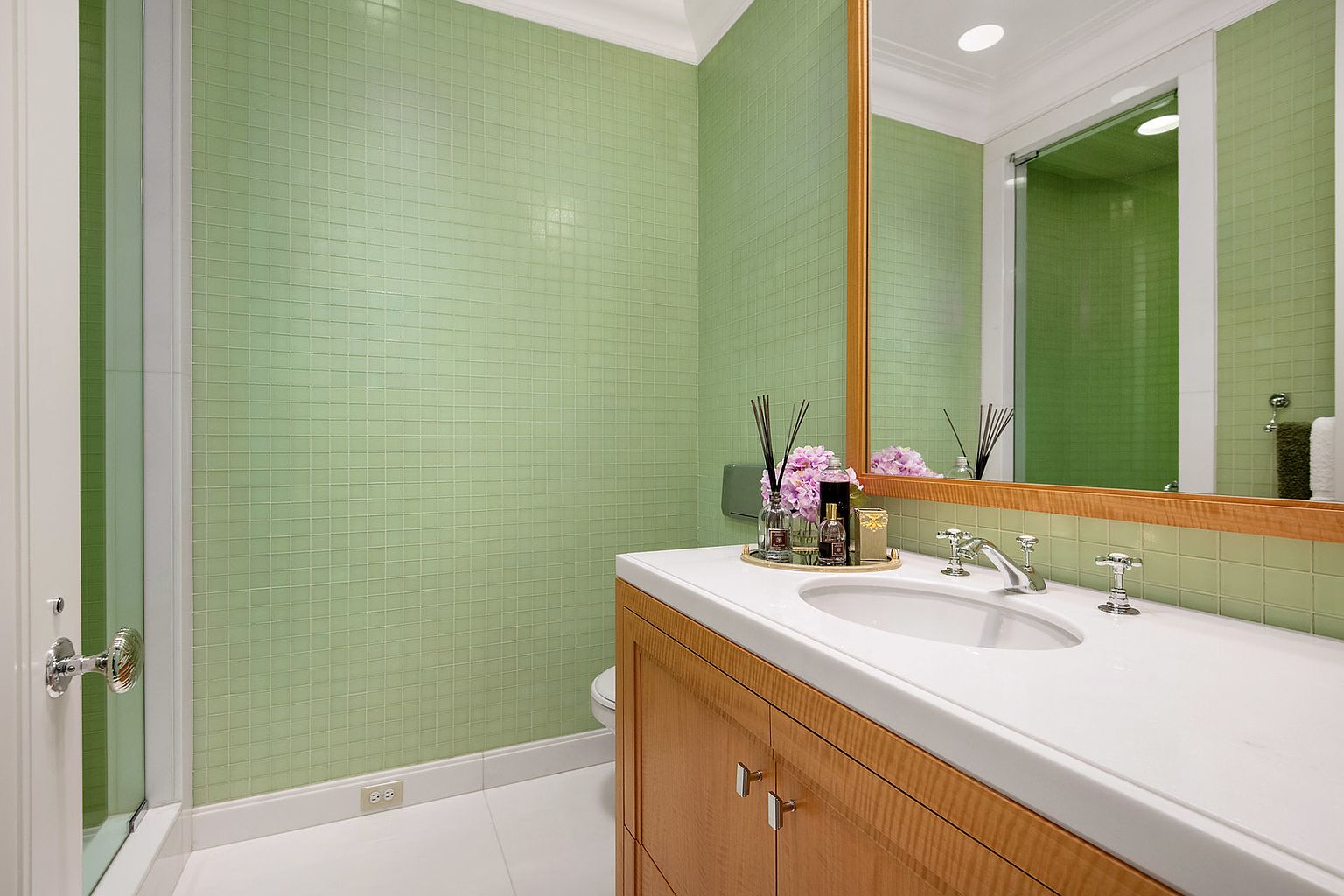 This is a bathroom featuring light green square tiles on the walls, a white countertop with an oval sink, and light wood cabinetry. A large mirror reflects the shower area, and the overall design is clean and bright. The perspective is from the doorway, showcasing the vanity and mirror.