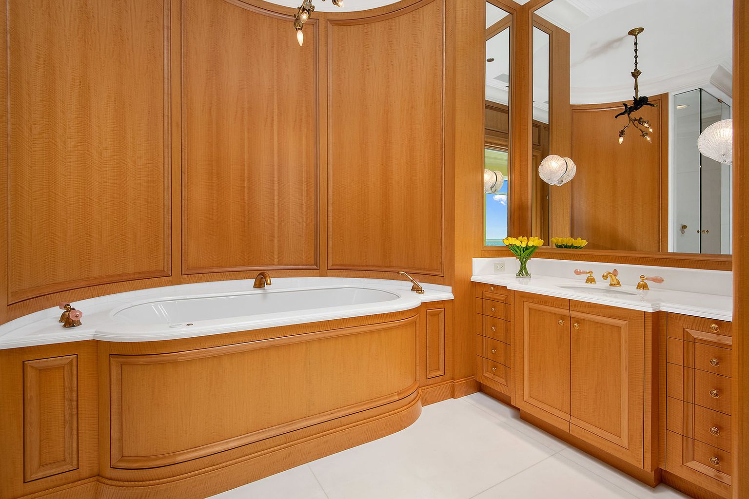 This is a luxurious primary bathroom featuring a curved bathtub with wood paneling and a white countertop. The vanity area includes wood cabinetry, a white countertop, and a large mirror. The room is well-lit with decorative lighting fixtures and has a clean, elegant aesthetic.