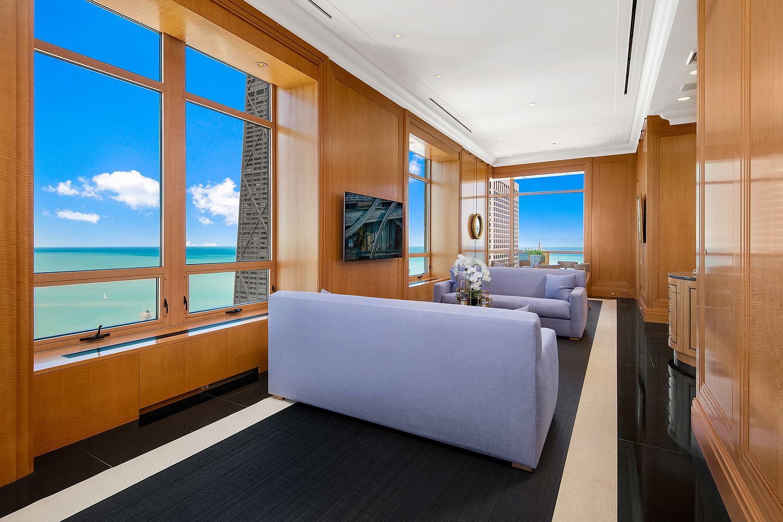 This is a luxurious living room featuring wood paneling, large windows with stunning water and city views, and elegant furnishings. The room is decorated in a sophisticated style with a neutral color palette, creating a serene and upscale atmosphere. The black and white striped rug adds a modern touch to the classic design.