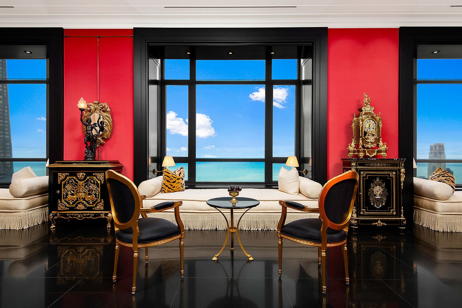 This opulent living room features a stunning view of the ocean through a large window, flanked by red walls and black trim. The room is furnished with ornate black and gold cabinets, elegant seating, and a small table, all set against a glossy black tile floor. The overall impression is one of luxury and sophistication.