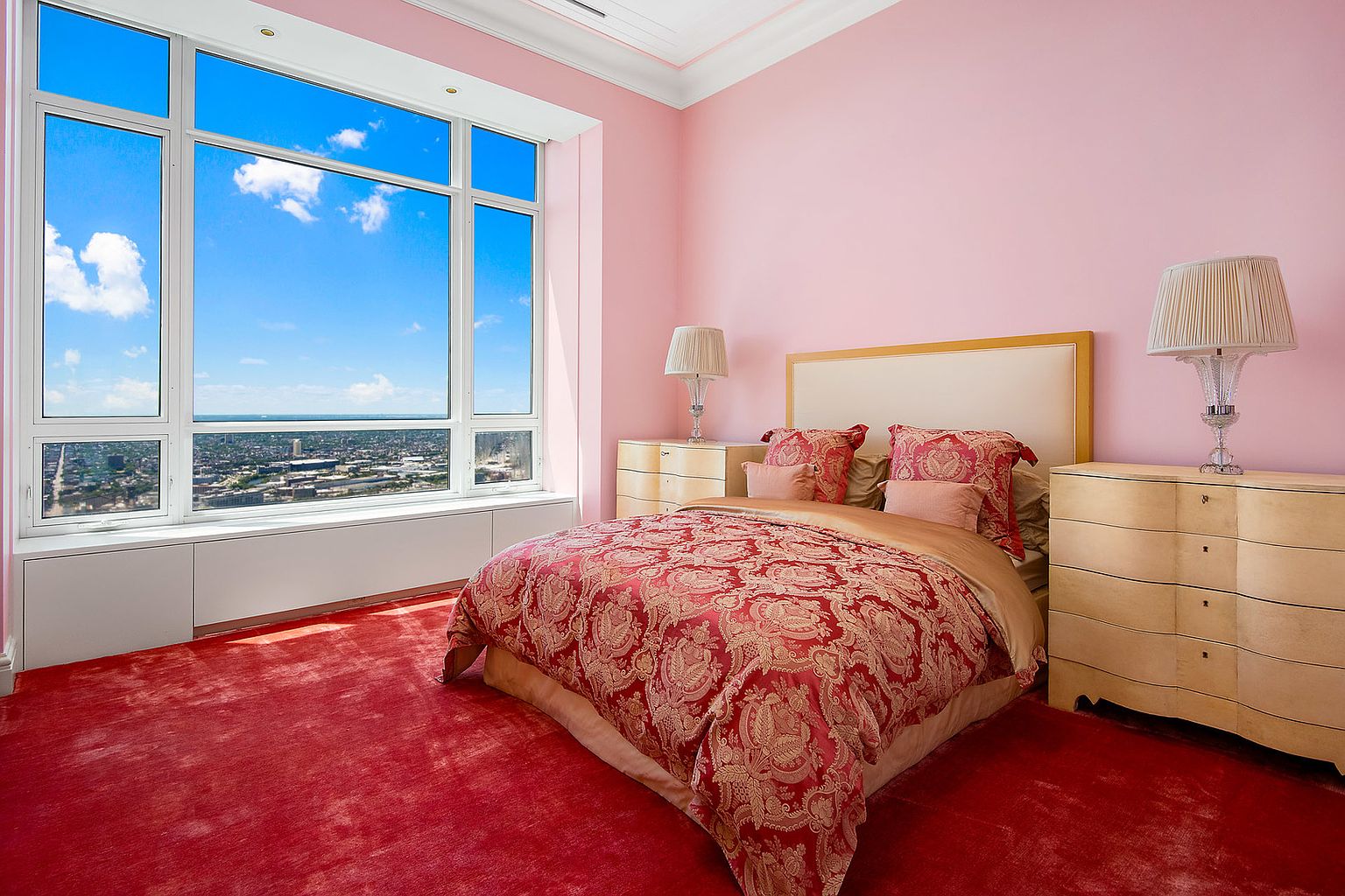 This is a primary bedroom featuring a large window with a city view, pink walls, and red carpeting. The bed is adorned with a patterned red and gold comforter and matching pillows. Two light-colored wooden nightstands flank the bed, each topped with a decorative lamp, creating a luxurious and comfortable atmosphere.