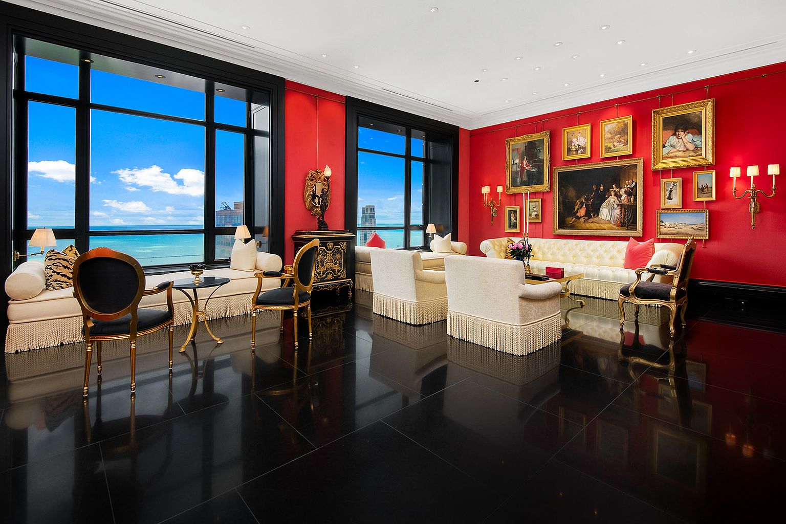 This opulent living room features a striking red wall adorned with classic paintings, complemented by elegant cream-colored sofas and armchairs with fringe detailing. Large windows offer a panoramic view of the ocean, while the glossy black tile floor reflects the room's luxurious furnishings. The overall impression is one of grandeur and sophistication.