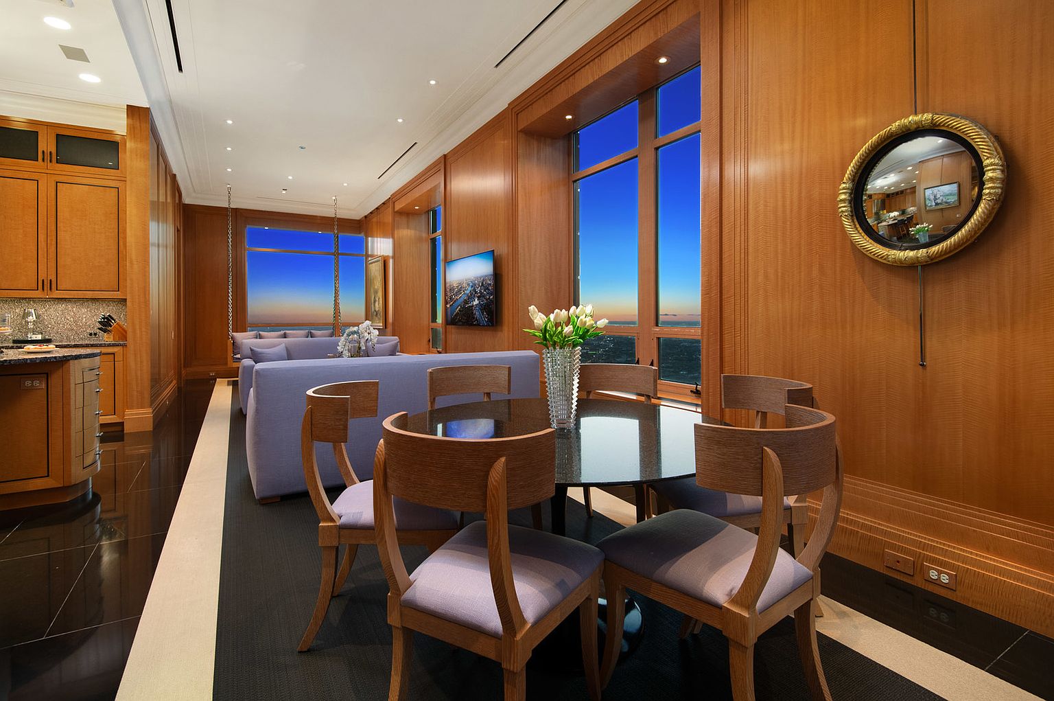This is an interior shot of a luxurious dining area. The room features wood-paneled walls, large windows with a city view, and a round dining table with modern chairs. A decorative mirror hangs on the wall, adding to the room's elegant ambiance.