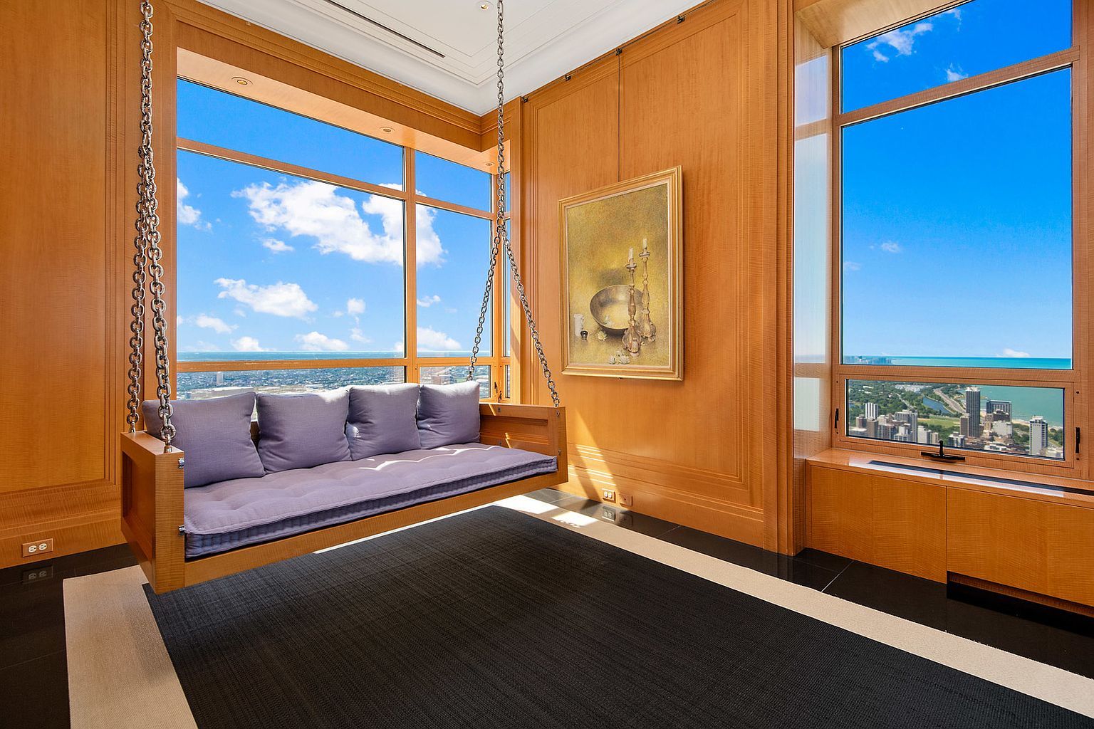 This interior shot showcases a luxurious living room with a unique hanging swing-bed positioned in front of a large window offering a stunning city view. The room features wood paneling, a piece of art, and a black and beige rug, creating a sophisticated and comfortable atmosphere. The perspective is from a low angle, emphasizing the height and grandeur of the space.