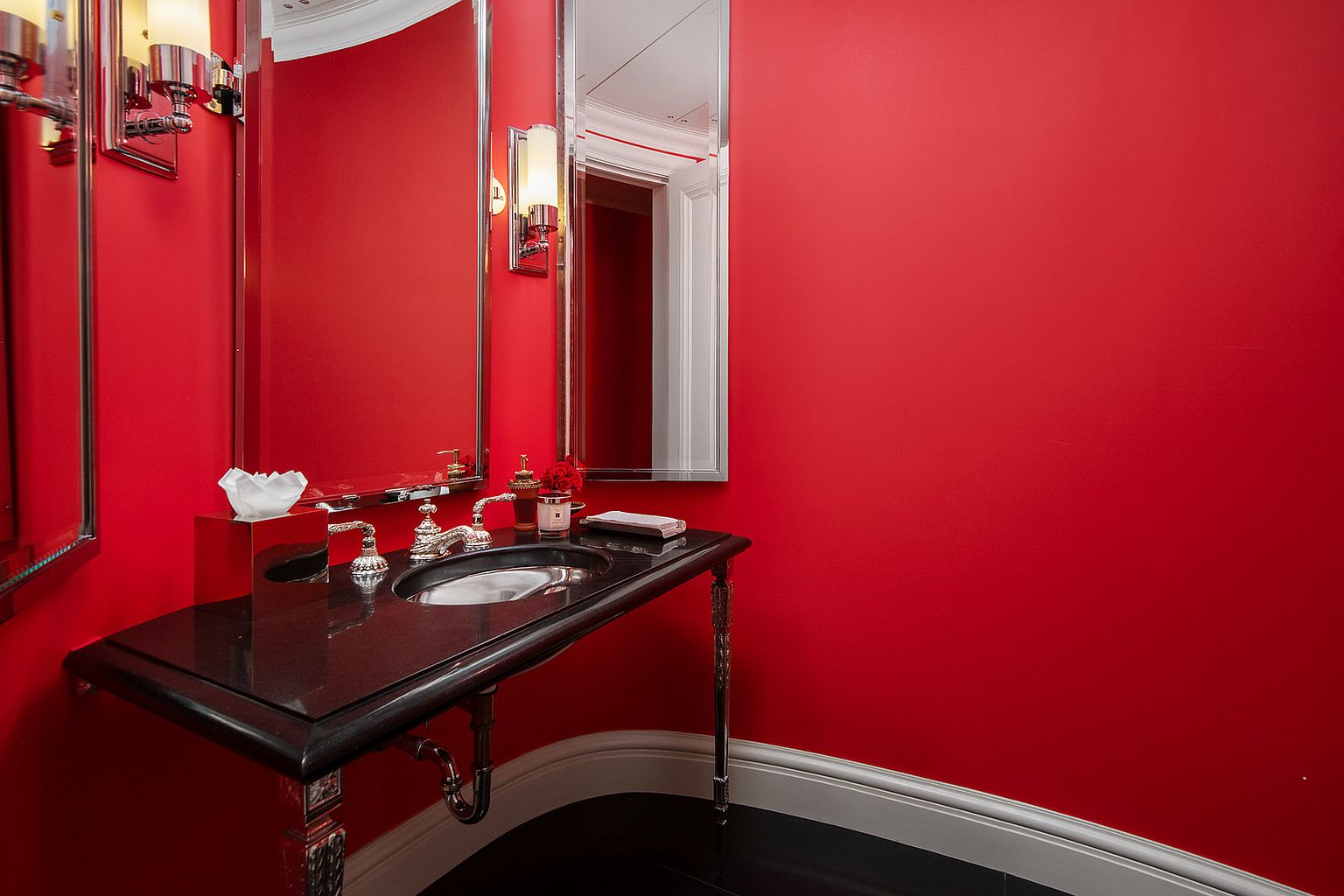 This is a striking guest bathroom featuring bold red walls, a black marble countertop vanity with ornate silver legs, and a silver-framed mirror. The room is illuminated by sconces, and the black flooring adds a touch of sophistication. The perspective is from a medium angle, showcasing the vanity and the vibrant color scheme.