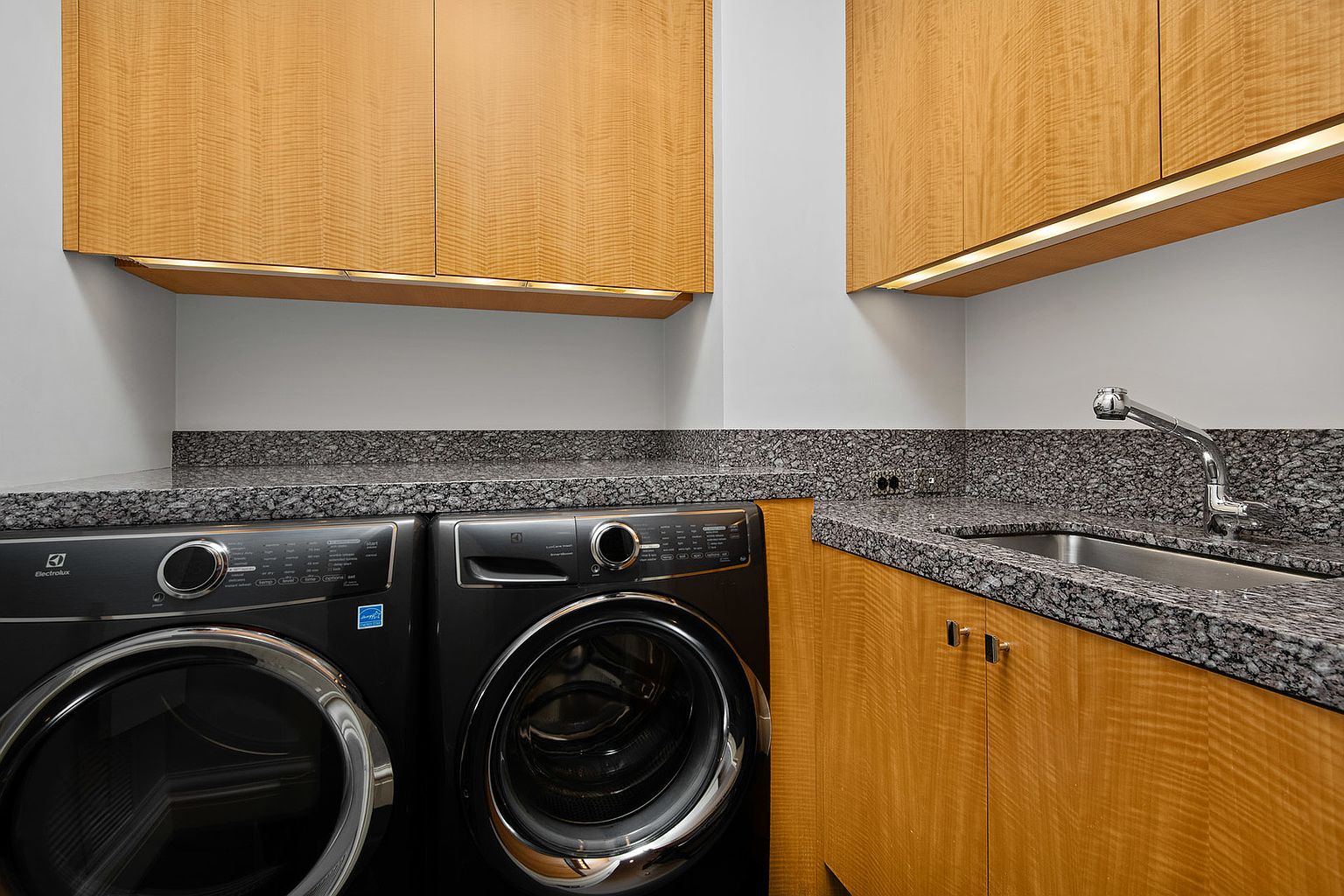 This is a well-appointed laundry room featuring modern, dark-colored front-loading washer and dryer units. The countertops are a speckled granite, complementing the light wood cabinetry above and below. A stainless steel sink and faucet are also present, creating a functional and stylish space.