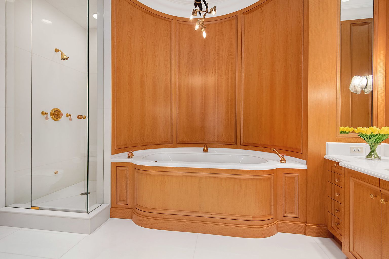 This is a luxurious primary bathroom featuring a large soaking tub surrounded by custom wood paneling. A glass-enclosed shower with gold fixtures is visible to the left, and a vanity with a white countertop and wood cabinetry is on the right. The white tile flooring and elegant lighting contribute to the bathroom's sophisticated and spa-like atmosphere.