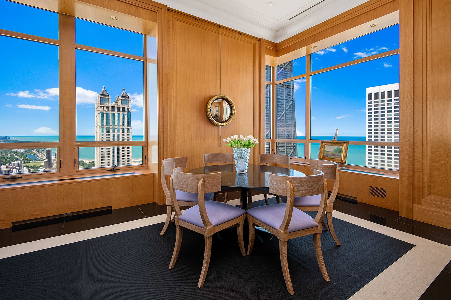 This is an elegant dining room featuring wood paneled walls and large windows offering stunning city and lake views. A round dark wood table is surrounded by six light wood chairs with lavender cushions. A round mirror hangs on the wall, and a vase of white tulips sits on the table, adding a touch of sophistication.