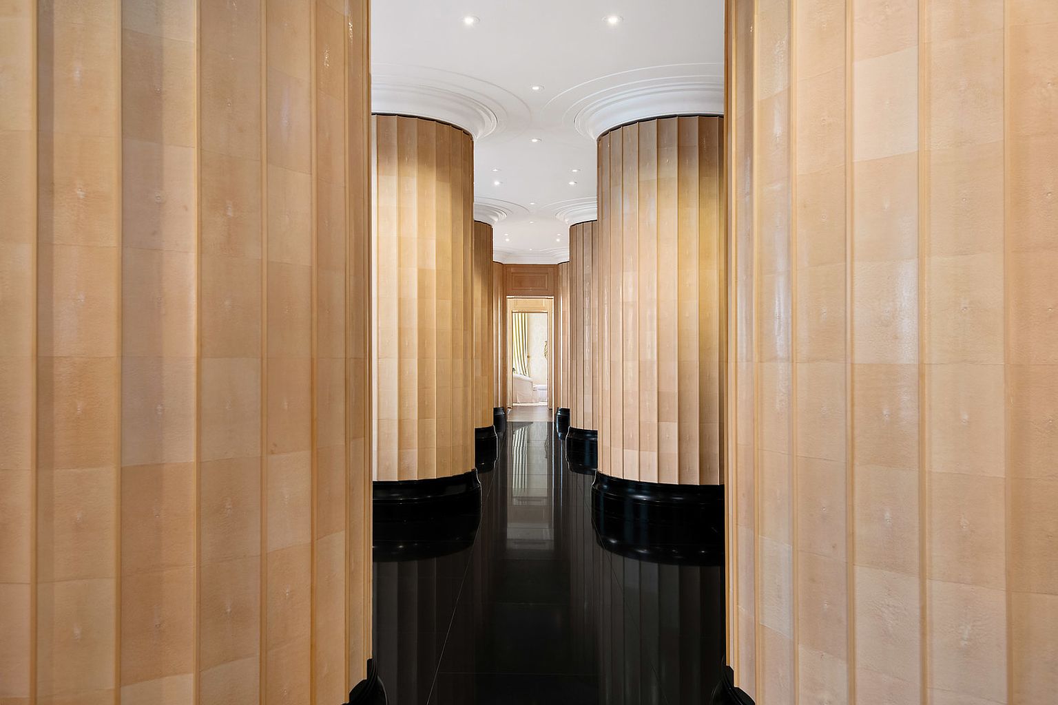 This interior shot showcases a luxurious hallway featuring fluted columns with black bases and a highly polished black floor that reflects the light. The hallway leads to a doorway at the end, suggesting a spacious and elegant home. The architectural details and high-end finishes create a sophisticated and upscale impression.