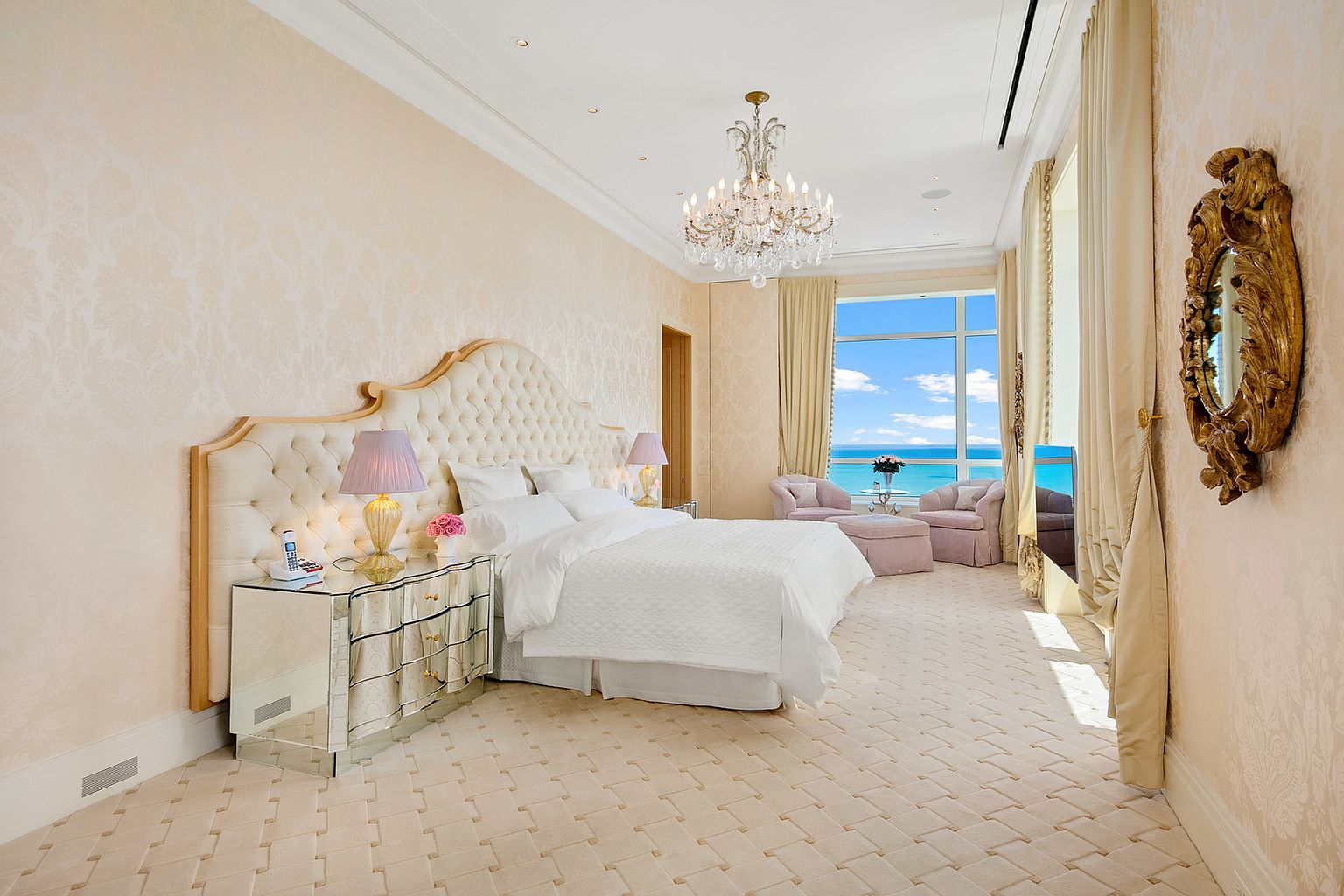 This is a luxurious primary bedroom featuring a large, tufted headboard and a white quilted bedspread. The room is decorated in a neutral color palette with patterned wallpaper and a crystal chandelier. Large windows offer stunning ocean views, and there is a seating area with plush chairs.