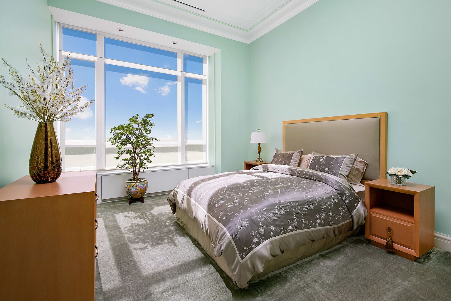 This is a serene primary bedroom featuring a large window with a view, a comfortable bed with a neutral-toned headboard and patterned bedding, and matching nightstands. The walls are painted in a calming light green, and the room is well-lit, creating a relaxing and inviting atmosphere. A dresser and decorative plants add to the room's appeal.