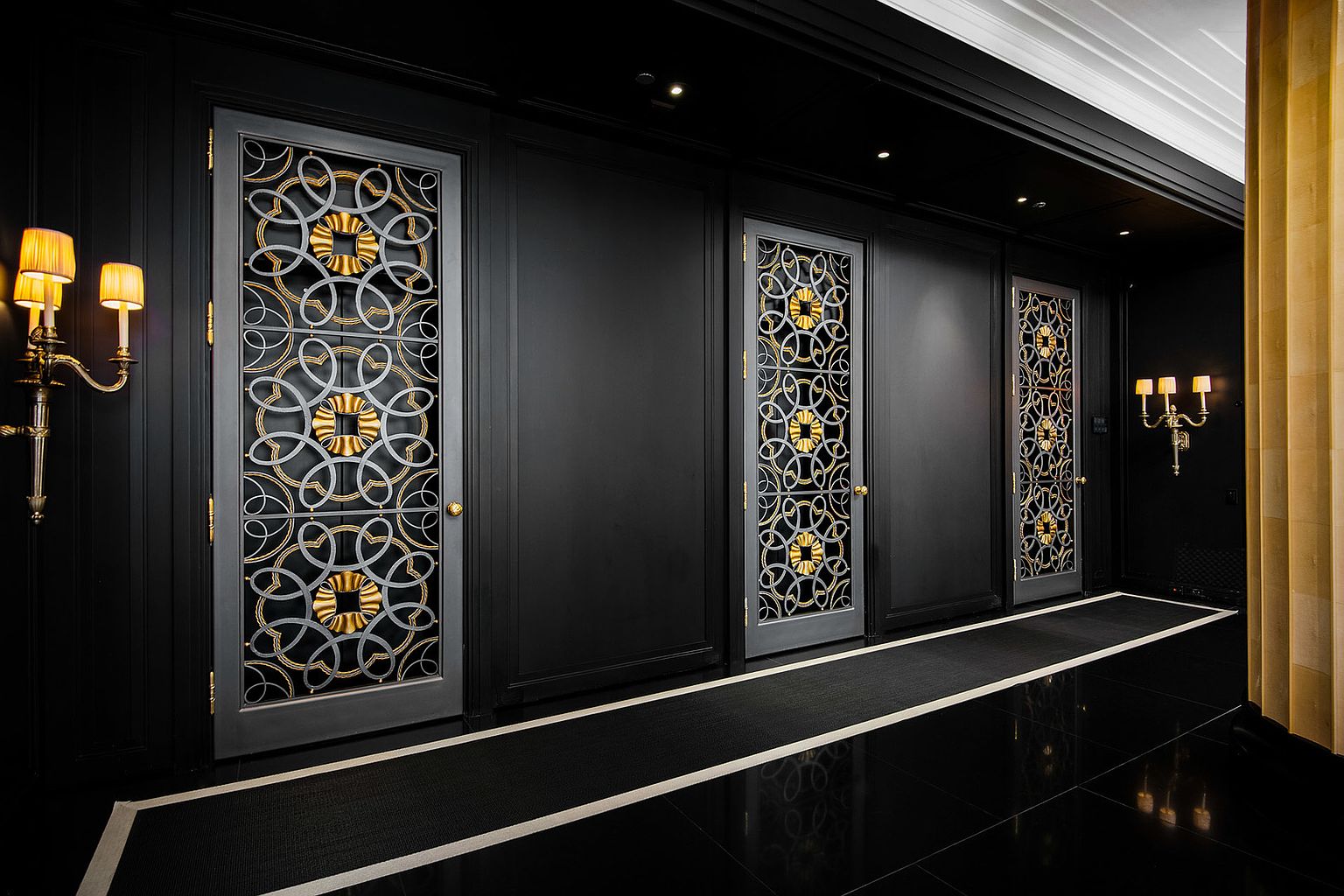 This is a luxurious hallway featuring black walls and a glossy black tiled floor with white accents. Three ornate doors with gold and silver detailing line the hallway, adding a touch of elegance. The lighting is warm and inviting, creating a sophisticated atmosphere.