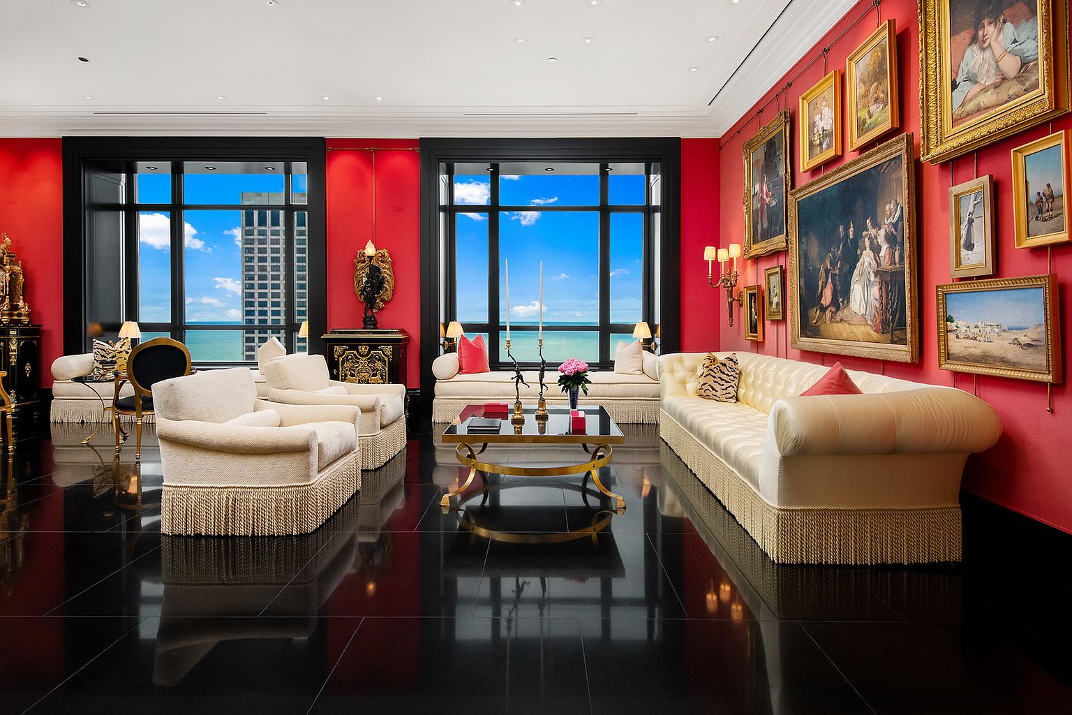 This opulent living room features a striking red wall adorned with classic paintings, complemented by elegant cream-colored sofas with fringe detailing. Large windows offer a stunning view of the cityscape and ocean, while the glossy black tile floor reflects the room's luxurious ambiance. The room exudes a sense of classic sophistication and grandeur.
