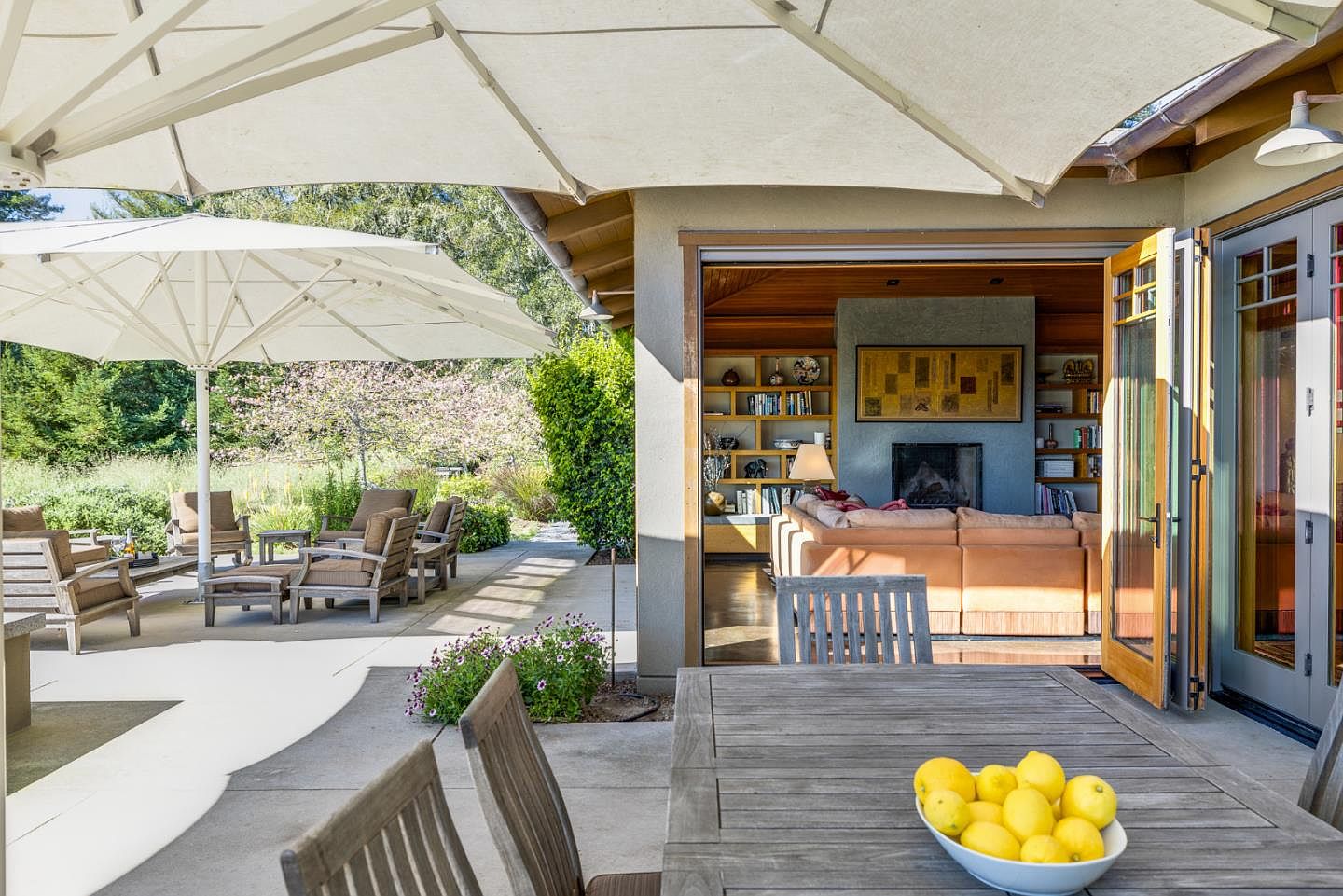 This inviting outdoor living space features a spacious concrete patio shaded by large umbrellas, seamlessly connecting to an indoor living area through wide-opening glass doors. The patio is furnished with comfortable wooden lounge seating and a dining table topped with a bowl of fresh lemons, creating a relaxed, resort-like atmosphere. The view looks from the dining area toward the cozy interior living room, which is anchored by a fireplace and built-in bookshelves, highlighting a perfect blend of indoor-outdoor California living.