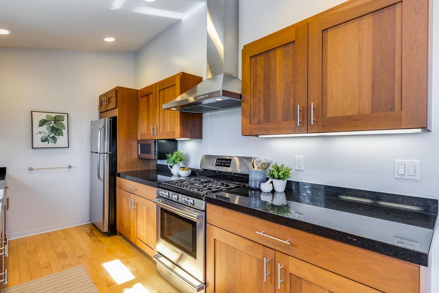 This well-lit kitchen features warm-toned wooden cabinetry paired with sleek black granite countertops and stainless steel appliances. The space includes a gas range with a modern vent hood, a built-in microwave, and a stainless steel refrigerator, all set against light-colored walls and hardwood flooring. The perspective is a straight-on, eye-level shot that highlights the functional layout and clean, contemporary aesthetic of the cooking area.