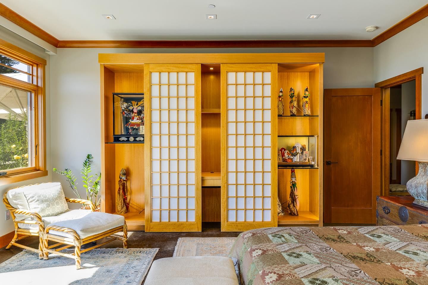 This serene primary bedroom features a distinct Japanese-inspired aesthetic, highlighted by a large, custom light-wood built-in unit with sliding shoji-style screens. The room is furnished with a comfortable rattan armchair, a patterned quilt bedspread, and decorative cultural artifacts displayed in illuminated shelving. The warm wood tones and soft natural light create a tranquil and sophisticated atmosphere.