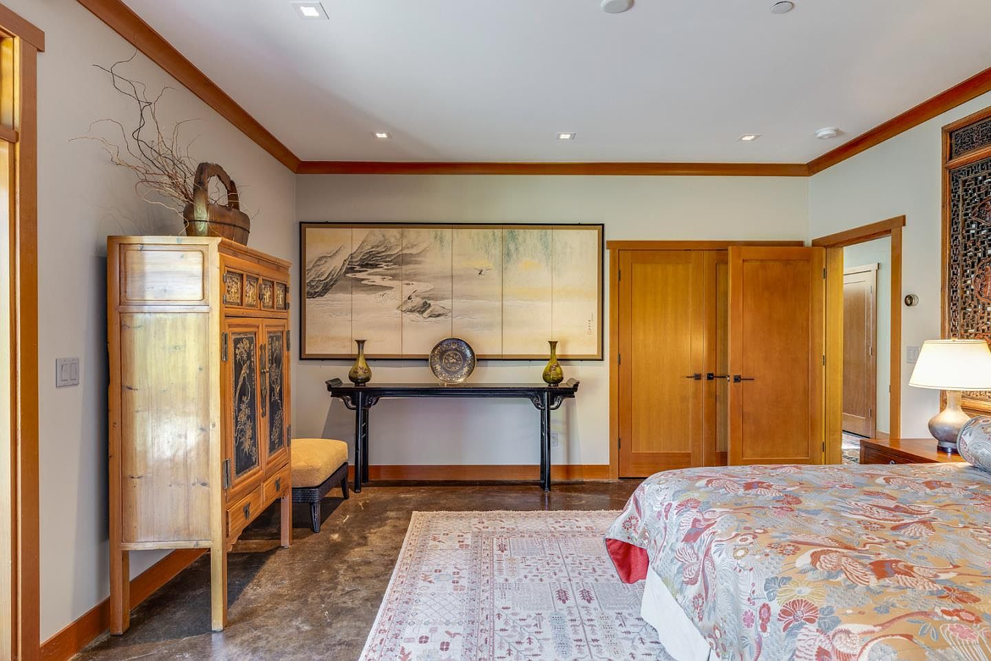 This primary bedroom features a sophisticated blend of Asian-inspired decor, including a large, ornate wooden armoire and a traditional landscape painting mounted above a sleek black console table. The room is anchored by a patterned bedspread and a large area rug, set against a backdrop of warm wood trim and polished concrete floors. The perspective is a wide, eye-level shot that captures the serene, curated aesthetic of the space.