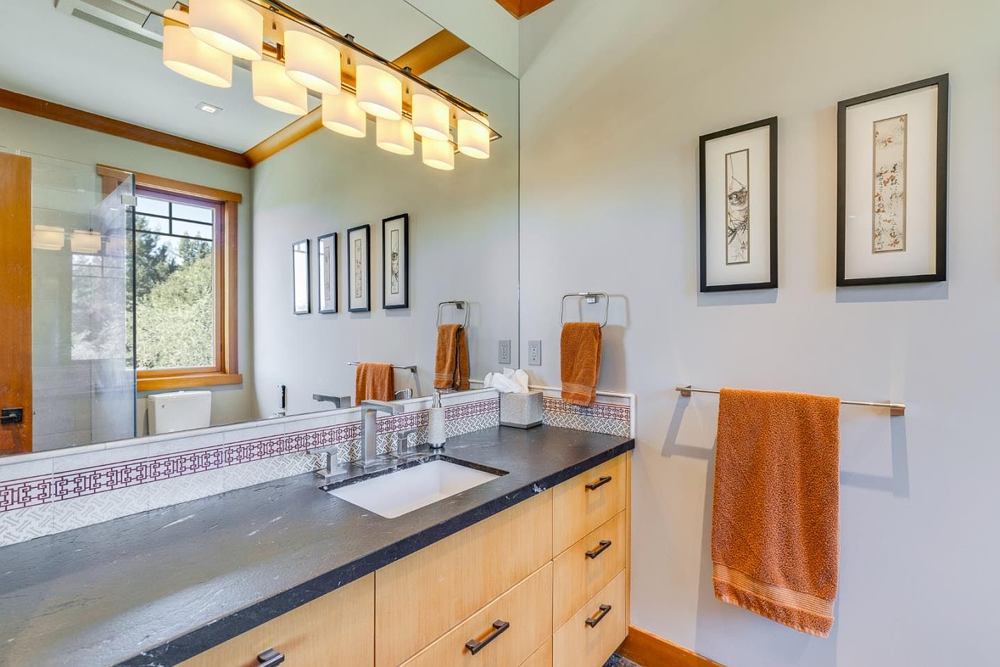 This well-appointed bathroom features a sleek, dark stone countertop paired with light wood cabinetry, creating a warm and modern aesthetic. A large mirror reflects the space, including a decorative tiled backsplash and a hanging orange towel, while framed artwork adds a personal touch to the light gray walls. The perspective is a straight-on shot of the vanity, highlighting the clean lines and functional design of the space.
