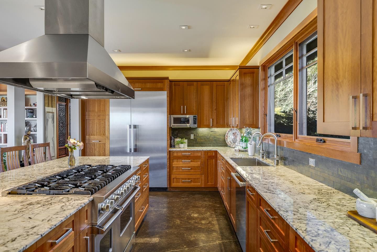This warm and inviting kitchen features rich cherry wood cabinetry paired with elegant granite countertops and a dark stone tile backsplash. The space is equipped with high-end stainless steel appliances, including a professional-grade range with a large hood and a built-in microwave. The perspective captures the functional layout from the island, highlighting the seamless flow between the cooking area and the sink, creating a sophisticated and practical culinary environment.