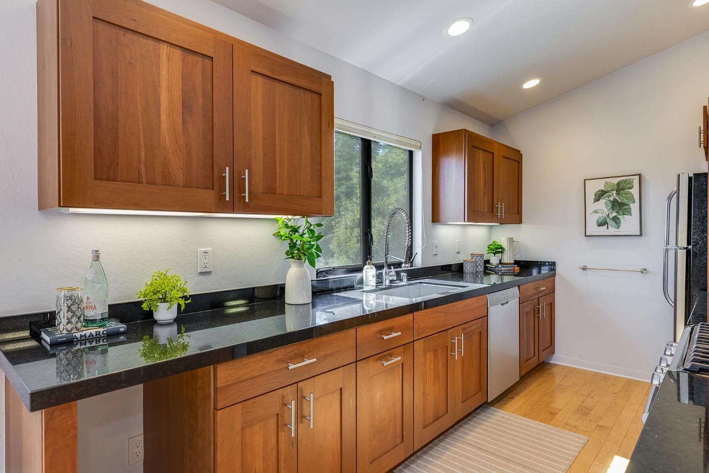 This well-lit kitchen features warm-toned wooden cabinetry paired with sleek black granite countertops, creating a sophisticated and inviting atmosphere. The space is equipped with stainless steel appliances, including a dishwasher and a faucet, while under-cabinet lighting highlights the workspace. A window above the sink brings in natural light, and the overall design balances traditional wood elements with modern finishes.