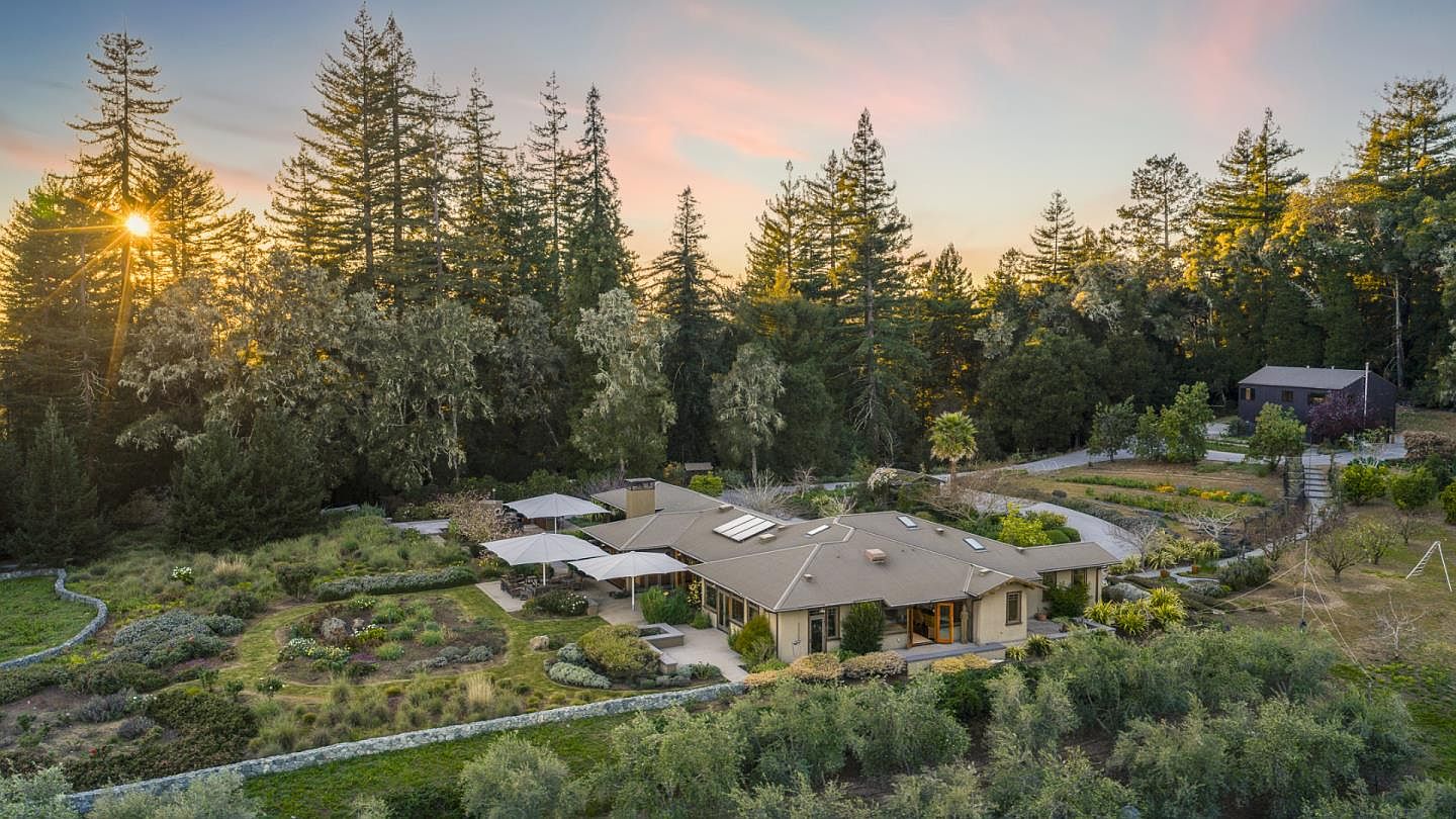 This stunning aerial perspective captures a sprawling, single-story residence nestled within a lush, wooded landscape at sunset. The property features extensive, meticulously landscaped gardens, a stone-walled courtyard, and a large patio area shaded by umbrellas, all set against a backdrop of towering redwood trees. The warm, golden light of the setting sun enhances the serene and private atmosphere of this expansive estate.