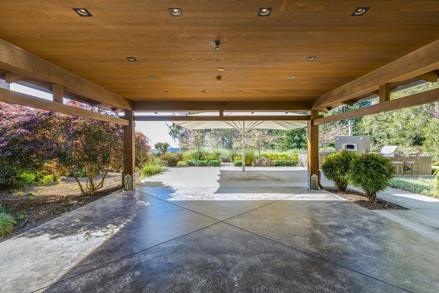 This expansive covered patio features a rich, wood-paneled ceiling with recessed lighting, providing a sophisticated transition between the indoor and outdoor living spaces. The concrete floor, marked with clean geometric lines, opens up to a sun-drenched backyard area complete with a large umbrella, an outdoor pizza oven, and lush landscaping. The perspective is framed by sturdy wooden support beams, creating a welcoming and airy atmosphere perfect for outdoor entertaining.