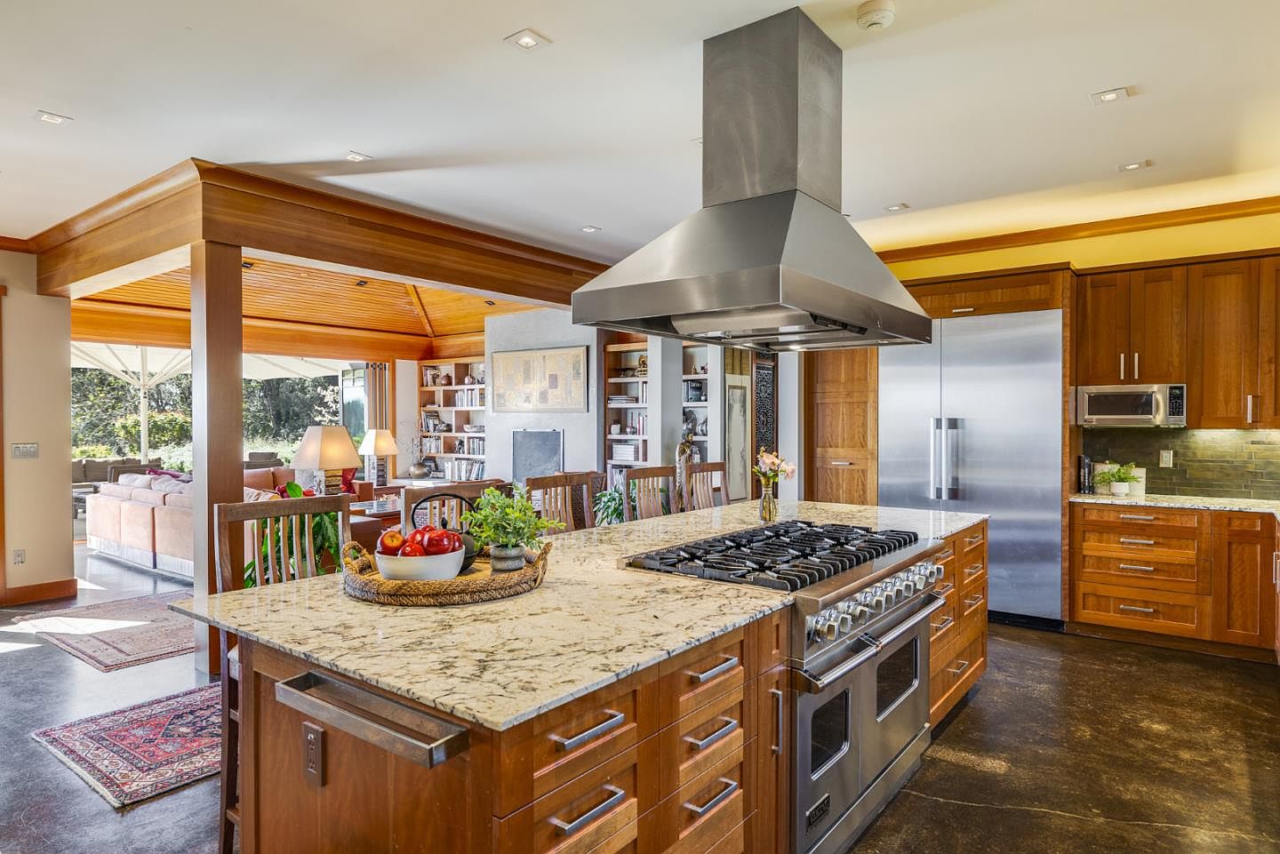 This spacious, open-concept kitchen features warm wood cabinetry, a large granite-topped island with a built-in gas range, and professional-grade stainless steel appliances. The perspective captures the seamless transition from the culinary workspace to a cozy, sun-drenched living area with vaulted wood-paneled ceilings. The overall aesthetic is a sophisticated blend of rustic warmth and modern functionality, perfect for entertaining.