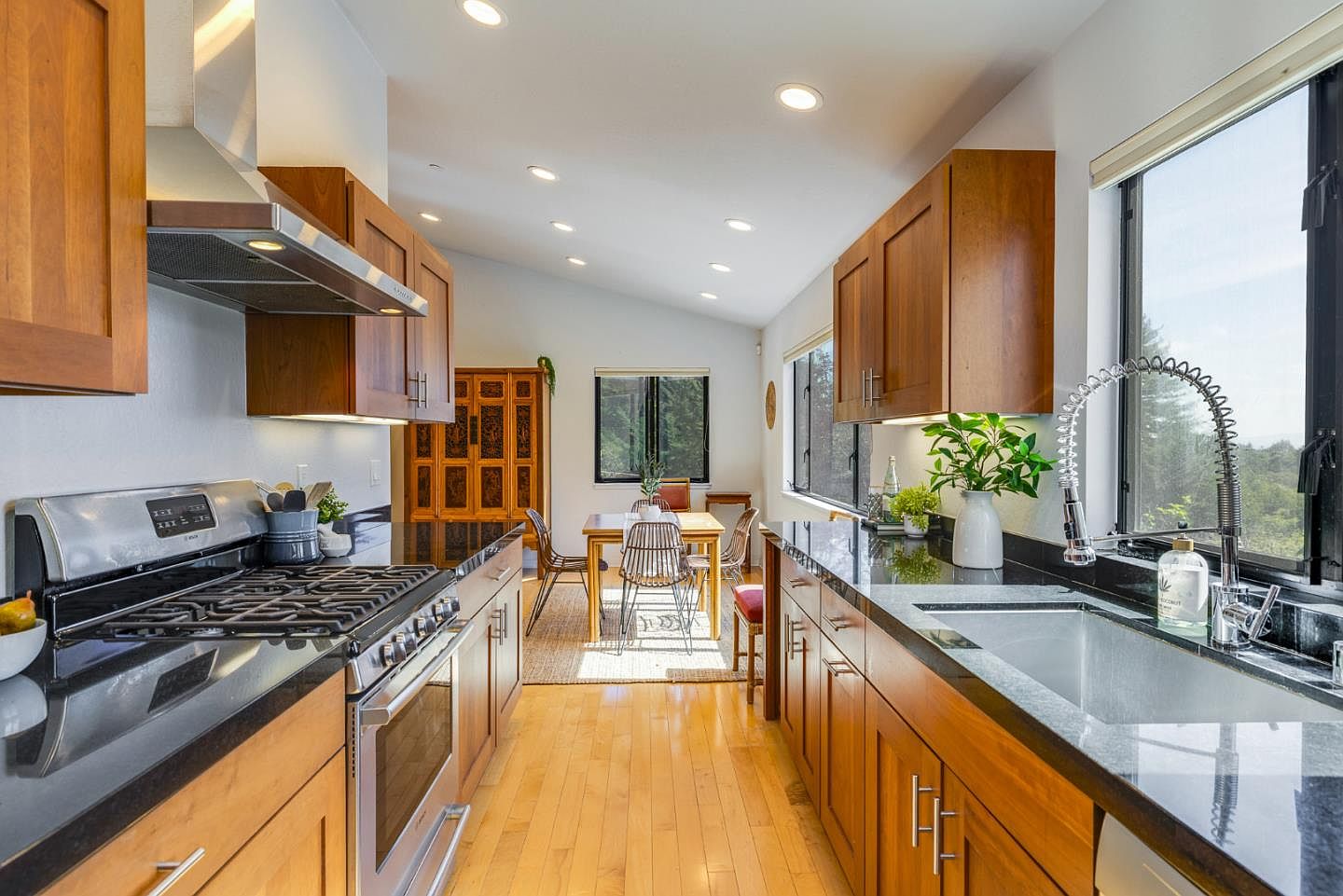 This bright and inviting kitchen features warm wood cabinetry paired with sleek black granite countertops and stainless steel appliances. The galley-style layout leads the eye toward a sunlit dining area in the background, creating a seamless flow between spaces. Large windows provide ample natural light and a scenic view, enhancing the overall airy and functional atmosphere of the home.