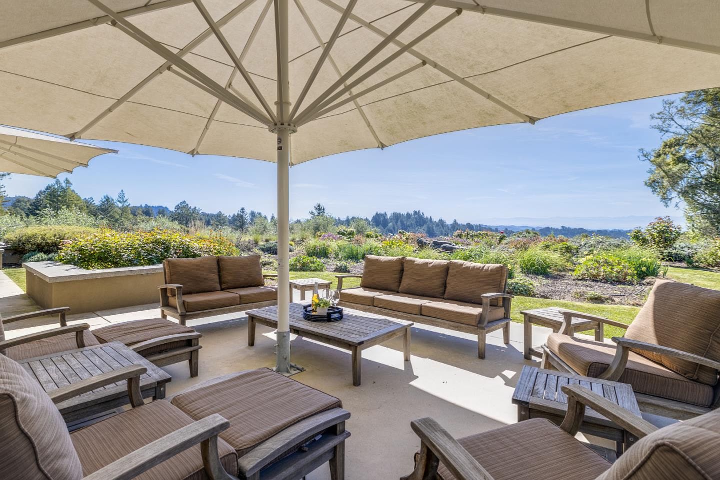 This inviting outdoor patio space features a comfortable seating arrangement with plush brown cushions on wooden frames, centered around a rustic coffee table. A large, expansive umbrella provides shade over the lounge area, which overlooks a beautifully landscaped garden and rolling hills in the distance. The scene captures a serene, high-end outdoor living environment perfect for relaxation and entertaining.