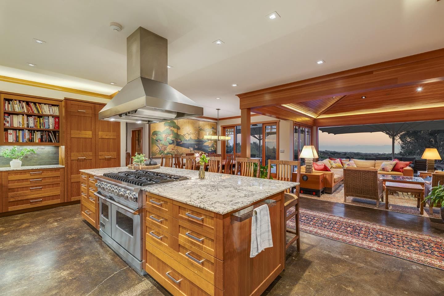 This spacious, open-concept kitchen features warm wood cabinetry, a large granite-topped island with a professional-grade range, and a striking stainless steel range hood. The kitchen seamlessly transitions into a cozy living area with expansive glass doors that reveal a stunning sunset view over the landscape. The combination of rustic wood tones, polished stone, and an open floor plan creates an inviting and luxurious atmosphere perfect for entertaining.